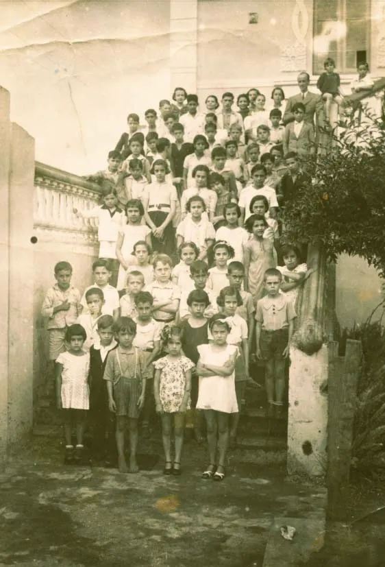 Grupo de crianças e adultos diante de uma escadaria, em fotografia antiga
